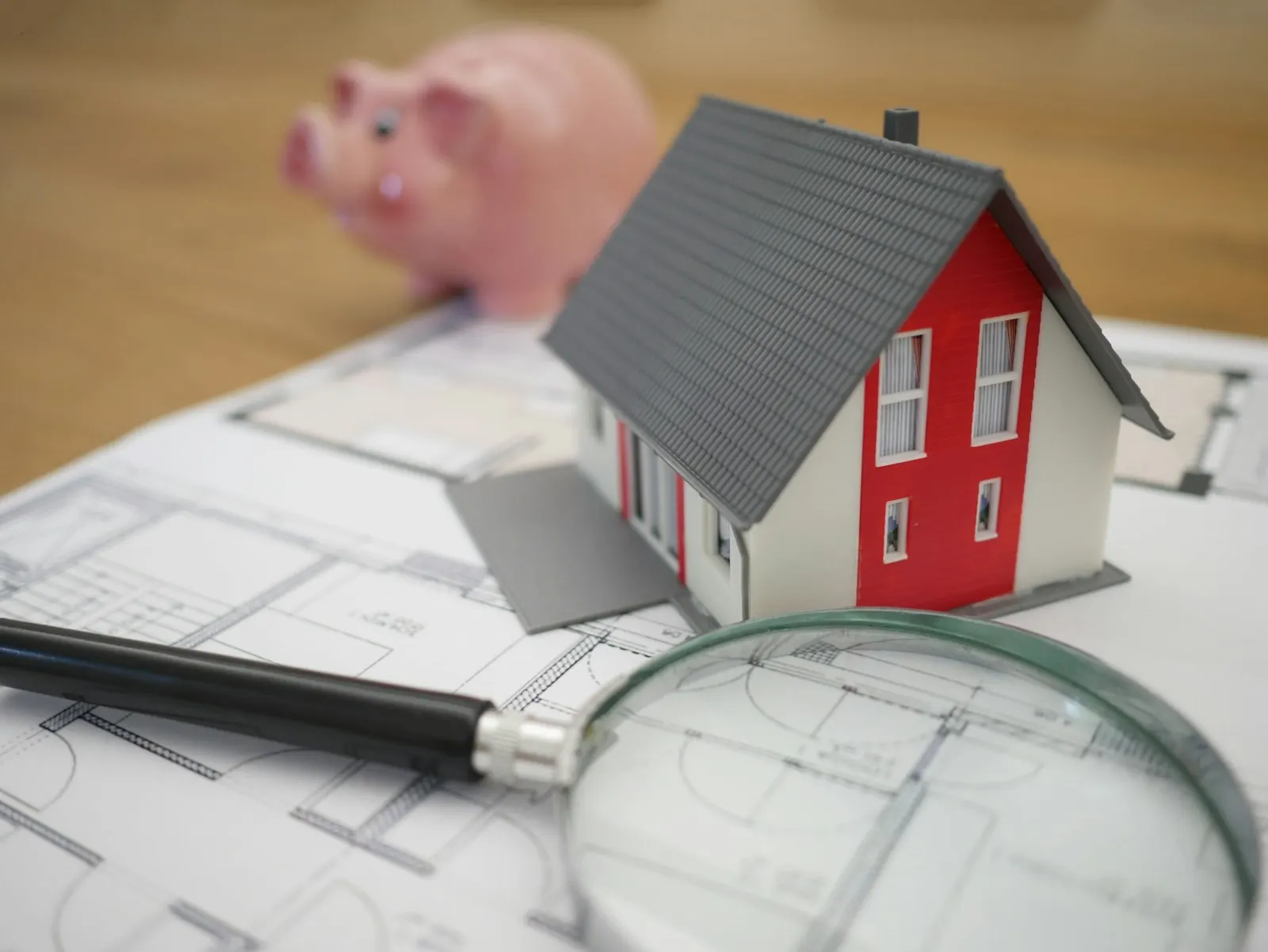 House model on top of architectural plans with a magnifying glass and a piggy bank